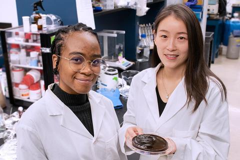 Two female scientists in a lab with a petri dish of a wet black substance