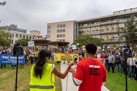 A photo of two young university workers shot from the back. They are standing on a stage, addressing a large crowd holding signs and placards with trade union logos