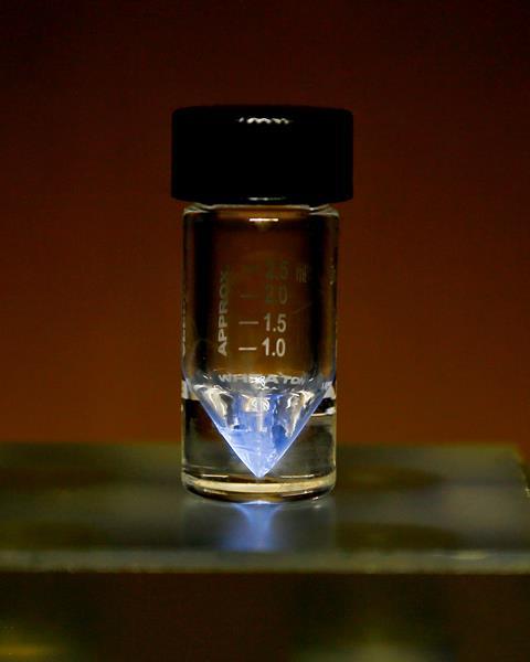 A photo showing a closeup of a small glass vial with a black plastic screw-on top. At the bottom of the pointed vial sits a small amount of blueish liquid that has a glowing shine to it.