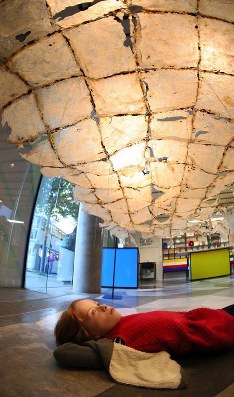 A girl at the plastic exhibition lying on the floor, looking up at a canopy made from plastics sheets suspended from the ceiling