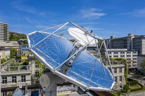 A photo showing a large parabolic dish lined with silvery mirrors. The system is sitting on an urban rooftop, a number of large period buildings can be seen in the background. It's a fine day with bright blue skies.