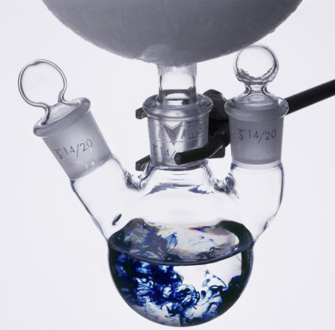 A photo of a three-neck round-bottom flask. Inside is a mostly clear liquid, with wisps of inky blue liquid swirling trough the middle