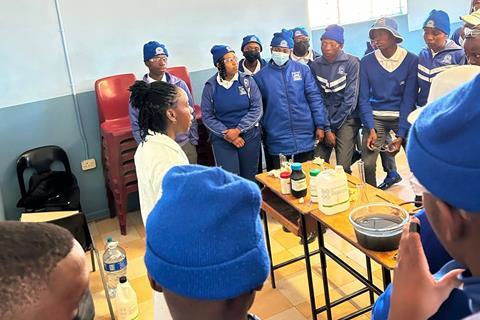 A group of students in Lesotho wearing a blue uniform with blue and white hats and coats in a chemistry lab with an instructor giving a demonstration.