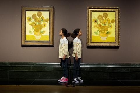 A photo of two versions of van Gogh's Sunflowers paintings hanging next to each other in a museum. Between them, standing back to back, are two Black girls, twins, wearing identical striped shirts but different coloured shoes (one pink, one grey).