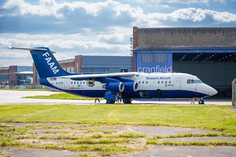 The FAAM research aircraft outside its hangar at Cranfield airport. It is a blue and white research aircraft parked on the apron in front of its hangar. There is green grass in front of the aircraft and blue sky with clouds in the background.