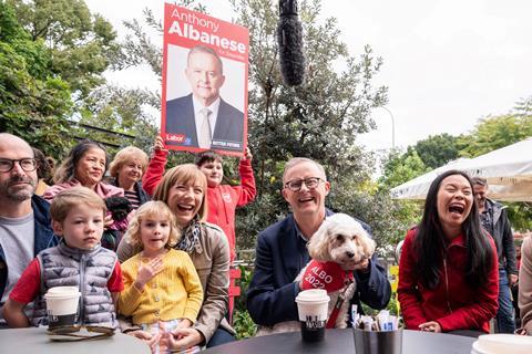 A white, middle-aged, bespectacled man seated at a table. He's laughing and holding a dog on his lap. There is a small crowd of people gathered to his right, one holding an election campaign poster.