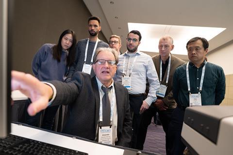 A man at a computer demonstrating to a group of conference attendees standing behind him