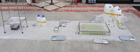 A photograph of an outdoor experimental setup laid out on a concrete surface. The setup consists of various scientific equipment, including plastic containers with liquid, tubes, metal plates and a small electronic device with wires