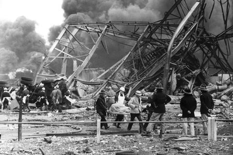 Black and white photo of people in safety gear pulling injured people form a destroyed building of twisted metal, rubble and smoke