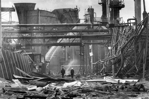 Black and white photo of twisted pipes and a ruined building there are jets of water being directed at some areas and people in safety gear walking through the wreckage