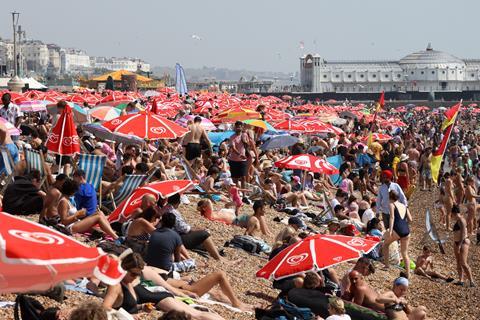 A dense crowd of people sunbathing on Brighton beach in the UK with lots of parasols and the pier and promenade in the background