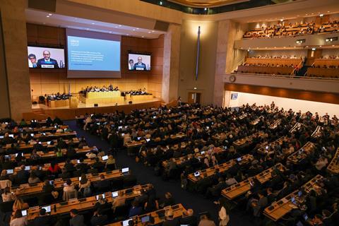 A large conference hall with people sitting at desks with screens and others on a stage