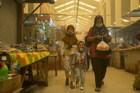 An image showing a mother with her two children walking through a market in Palangka Raya; they are wearing face masks as the space is filled with smoke produced by a wildfire, which also obstructs visibility