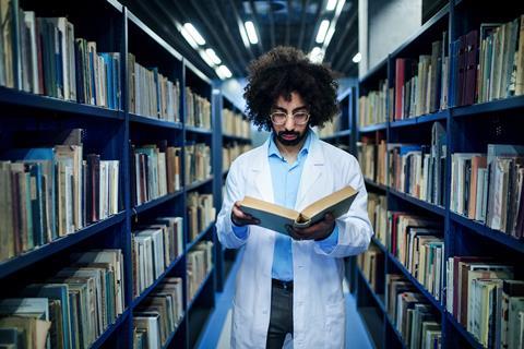 A man wearing a lab coat reading a book between the shelves of a library