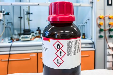 A brown bottle of solvent with hazard pictograms for flammable, harmful and harmful to the environment in a lab with kit in a fume cupboard behind