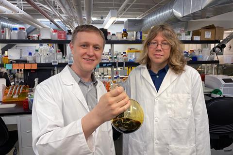 Two scientists in lab coats in a modern lab. One is holding a glass flask with a dark liquid inside.