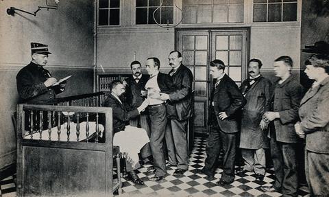 A 19th century black and white photo of men in suits queueing to be vaccinated