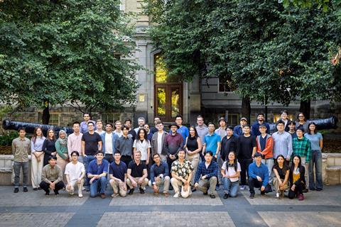 A team of people organised into rows for a group photo outside a grand building with trees and two old canons