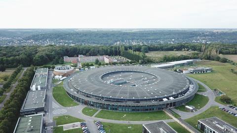 Aerial view of a large circular scientific facility surrounded by buildings, trees, and parking areas in a semi-rural landscape