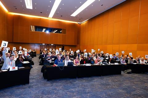 A photograph showing council members voting at the Iupac general assembly in Malaysia in July 2025. The council members are seated in a large auditorium, many are holding numbered cards in the air.