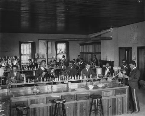 A black-and-white photo of a busy, early 20th century lab. Along wooden benches, men - wearing suits - carry out experiments, holding glassware or pipetting reagents