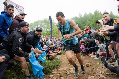 A male athlete running up a rough path on a steep hill through a crowd cheering him on