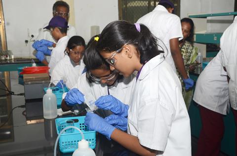 A pair of high school students in a practical chemistry class carefully pouring crystals into a funnel