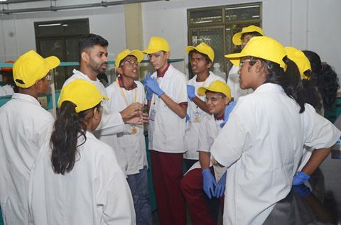 A scientist speaking to a group of visually impaired high school students in a lab. They are wearing yellow caps, lab coats and safety goggles
