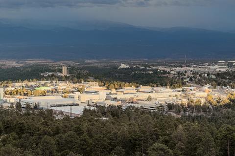 An aerial view of Los Alamos National Laboratory in the USA. There a number of low-rise office-type buildings surrounded by forest with cloud-topped mountains in the distance
